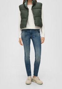 Olijfgroene gewatteerde bodywarmer met een hoge kraag, gecombineerd met een wit shirt met lange mouwen en blauwe skinny jeans. Beige sneakers maken de look compleet.