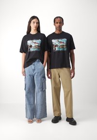 Black graphic t-shirts feature a colorful illustration. The female model wears light blue wide-leg jeans, while the male model has beige trousers.