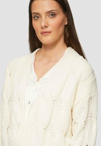 Kabelstrick-Cardigan in Creme, mit einem tiefen V-Ausschnitt und strukturiertem Muster. Wird über einer weißen Bluse mit Knopfdetails getragen.