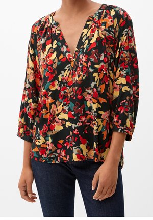Woman wearing a multicolored floral blouse with 3/4 sleeves and dark blue jeans, standing against a white background.