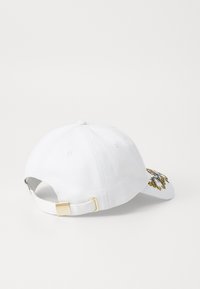 Cappellino da baseball bianco con design ricamato in oro e nero, fascia regolabile con accessori in oro e tessuto di cotone testurizzato.