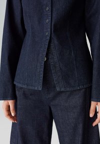 Dunkle Denimjacke mit tailliertem Schnitt, fünf Knöpfen, spitzem Kragen und seitlichen Schlitzen, kombiniert mit passenden dunklen Denimhosen.
