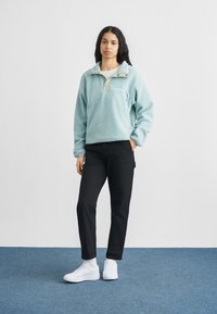 Νεαρή γυναίκα στέκεται με το ένα χέρι στην τσέπη φορώντας ανοιχτό μπλε φλις πουλόβερ, μαύρο παντελόνι και λευκά αθλητικά παπούτσια σε μπλε δάπεδο.