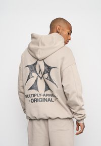 Felpa beige con un grande design grafico nero sul retro, con il testo "MULTIPLY.APPAREL ORIGINAL" e una vestibilità rilassata.