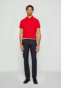 Hombre vestido con una camiseta polo roja, jeans oscuros y zapatos negros, de pie contra un fondo gris claro liso.