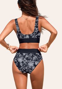 Bikini s vysokým pasem v černé látce s bílým květinovým vzorem, se sklopným zády a širokými ramínky pro podporu. Měkká textura s elastickým páskem.