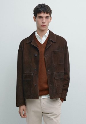 Bruine suède jas met een kraag, knoopsluiting en twee steekzakken aan de voorkant. Draagt over een bruine trui en een wit overhemd, gecombineerd met lichte broek.