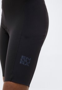 Detailní záběr černých cyklistických šortek na osobě s nápisem "ENDURA" vytištěným na stehně.