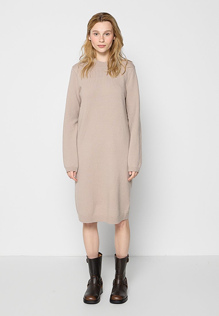 Hellbeige gestricktes Kleid mit langen Ärmeln und rundem Halsausschnitt, das knapp unter das Knie reicht, kombiniert mit dunkelbraunen Stiefeletten.