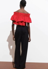 Top rosso scollato con strati arricciati, abbinato a pantaloni neri a gamba larga e sandali piatti, visto da dietro su uno sfondo bianco.