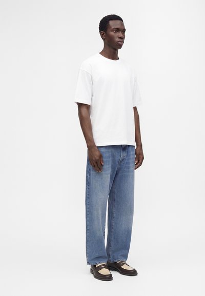 Jeune homme portant un t-shirt blanc uni à manches courtes, un jean bleu ample et des mocassins marron et beige, debout devant un fond blanc.