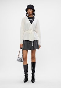 Světlý krémový zavinovací kardigan s vázáním, doplněný o skládací šedou sukni, černé lesklé holínky po kolena a stříbrnou kabelku.