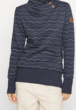 Granatowy sweter w długim rękawem z wzorem w zygzak, z golfem i guzikami, noszony z beżowymi spodniami i złotymi pierścionkami na palcach.