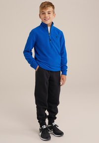 Pullover in pile blu con colletto zip, abbinato a pantaloni sportivi neri. L'outfit presenta un design semplice e una vestibilità rilassata per il massimo comfort.