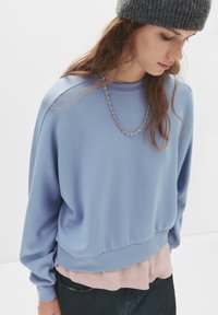 Felpa azzurra chiara con vestibilità rilassata, dettagli a costine sul collo e sui polsini, indossata sopra una camicia rosa morbida. Collana in argento come accento.