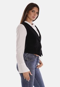 Gilet in velluto nero con un design aderente, due bottoni e tasche laterali, abbinato a una camicia bianca con bottoni e jeans blu.