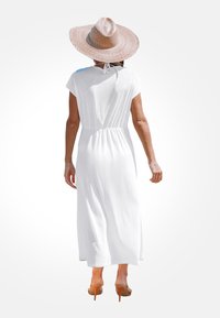 Robe maxi légère et blanche avec une taille smockée, des manches courtes et une jupe fluide. Portée avec un chapeau de paille à large bord et des sandales à talons.