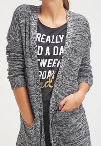 Szary dzianinowy kardigan o luźnym kroju, z długimi rękawami, otwartym przodem i dwoma przednimi kieszeniami, nałożony na czarny t-shirt z białym napisem.