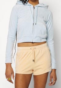 Person med en ljusblå kortärmad hoodie och orange shorts, med långa svarta flätor och ringar på fingrarna, mot en enkel bakgrund.