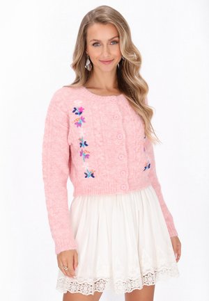 Rosa gestrickte, kurz geschnittene Strickjacke mit floralen Stickereien, Knopfverschluss und gerippten Bündchen. Kombiniert mit einem weißen Rock mit Spitzenbesatz.