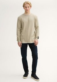 Beige langärmliges gestreiftes Hemd mit weißem Halsausschnitt, kombiniert mit dunkelblauen Jeans und schwarzen Sneakers. Auffälliges Detail mit Patch am Handgelenk.