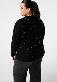 Zwarte trui met ronde parelaccenten, met een losse pasvorm en lange mouwen. Gecombineerd met donkergrijze wijde broekspijpen.