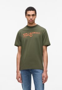 Mladý muž v olivově zeleném tričku s logem Karl Lagerfeld Jeans a oranžovým textem, ve modrých džínách, stojící na bílém pozadí.