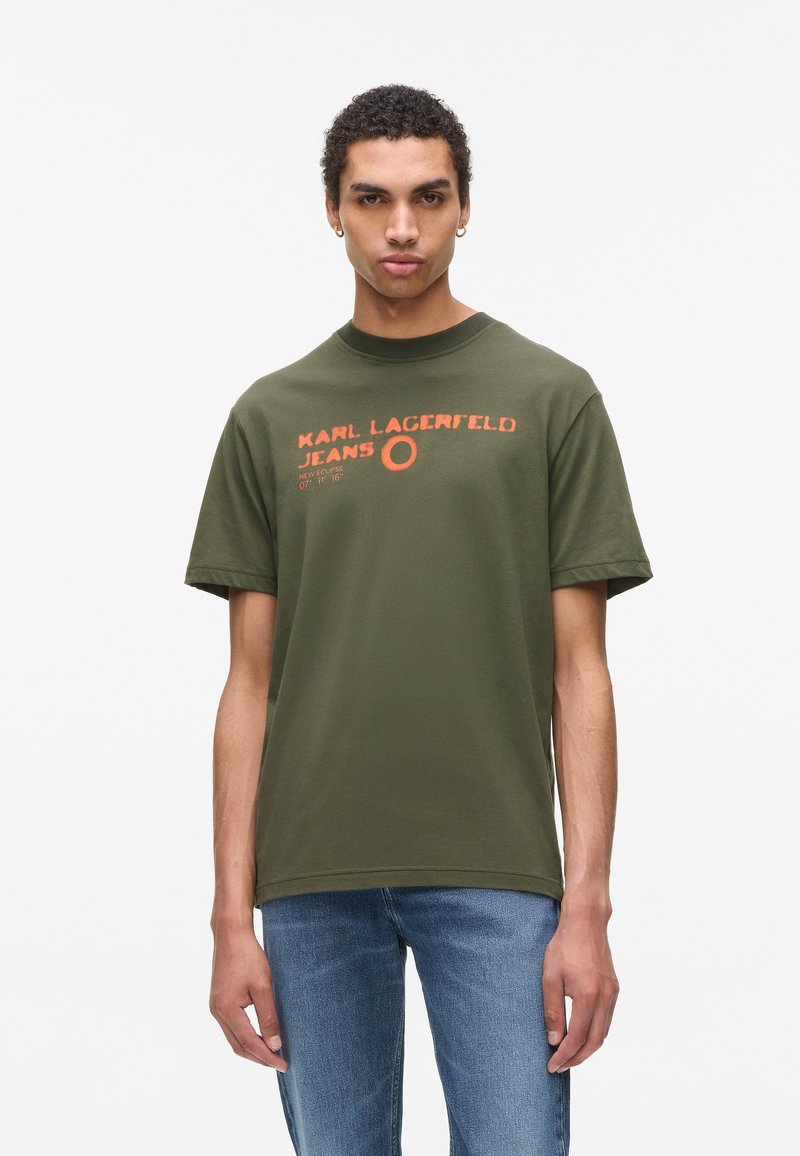 Jaunas vyras, vilkintis alyvuogių žalios spalvos Karl Lagerfeld džinsus, t-shirt su oranžiniu tekstu ir mėlynomis džinsomis, stovi prieš baltą foną.