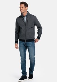 Graue Zip-Up-Jacke mit leichter Textur, gerippten Bündchen und einer Brusttasche, kombiniert mit Jeans und schwarzen Sneakers.