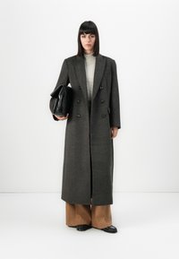 Personne portant un long manteau noir gris à double boutonnière, un col roulé rayé, un large pantalon marron, des chaussures noires, tenant un grand sac noir.