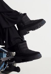 Black insulated boots with quilted upper, plush trim, textured sole, and wide footbed. Positioned on a wheelchair footrest.
