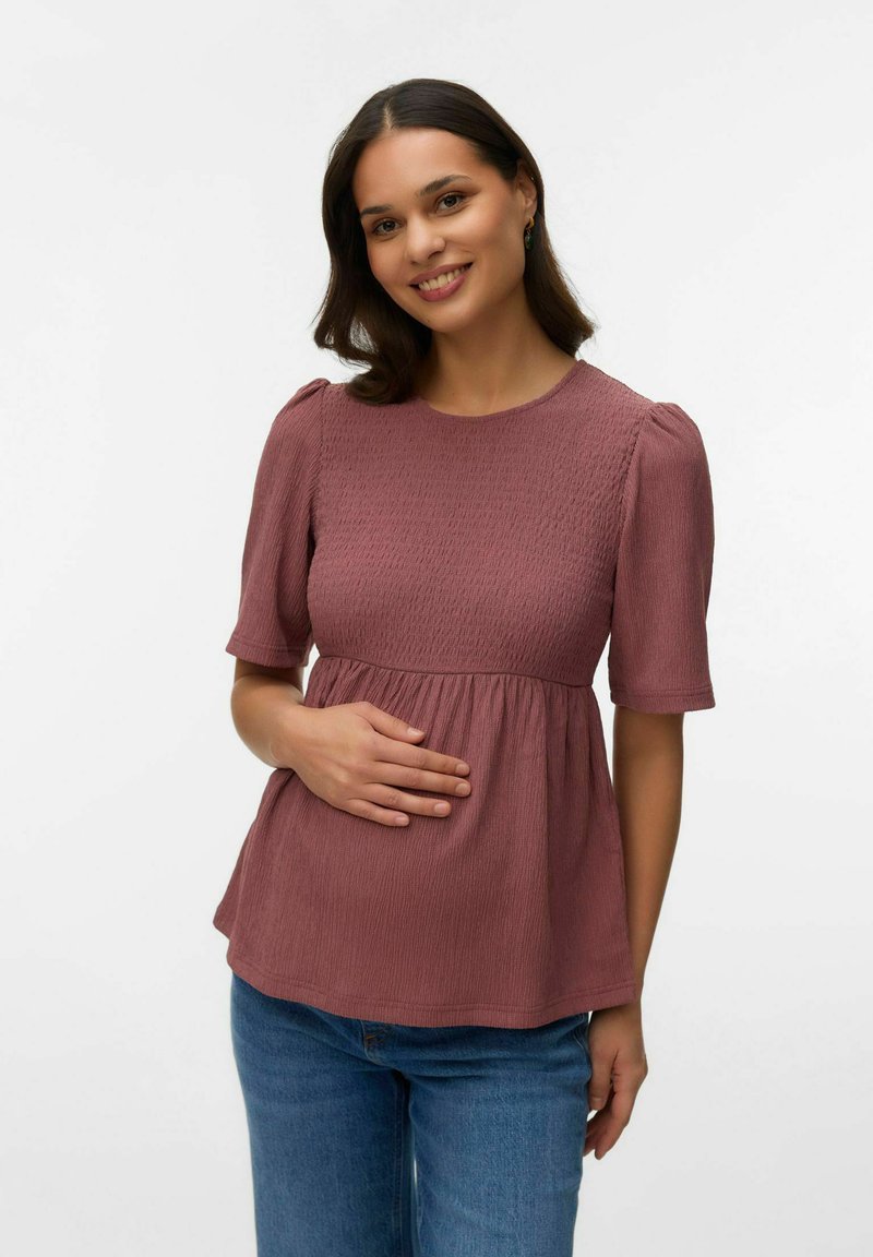 Naeratav tumejuukseline naine, kes kannab maasikaroosat tekstuuriga toppi ja siniseid pükse, hoides õrnalt oma kõhtu, seistes tavalise valge tausta ees.