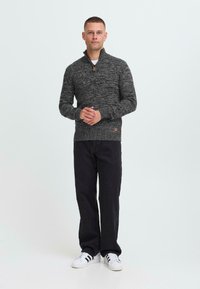 Uomo in piedi, che indossa un maglione grigio scuro a maglia con zip a quarti, pantaloni neri e scarpe bianche con strisce nere, su uno sfondo neutro.