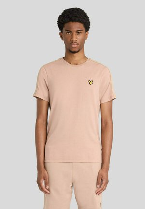Giovane uomo con capelli corti e ricci che indossa una maglietta beige chiaro e pantaloni coordinati, in piedi davanti a uno sfondo grigio uniforme.