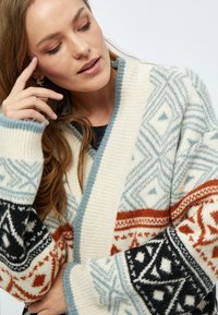 Cardigan épais avec une base blanc cassé et des motifs géométriques en bleu, noir et rouille. Texture tricotée douce et manches larges.