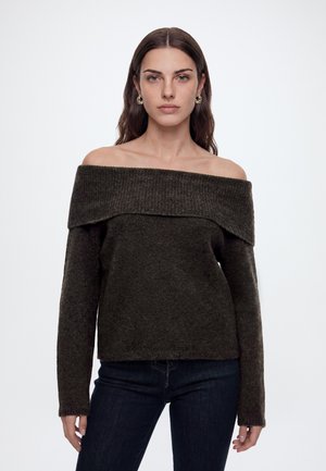 Femme aux longs cheveux bruns portant un pull noir épaules dénudées et un jean foncé, debout devant un fond blanc uni.