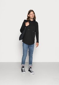 Sort langærmet hoodie, blå slim-fit denim jeans og sorte high-top sneakers med hvide såler, stående mod en ensfarvet hvid baggrund.