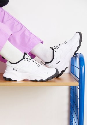 Jambes en pantalon violet et chaussettes blanches portant des baskets Nike blanches et noires reposant sur une étagère en bois avec un cadre métallique bleu.