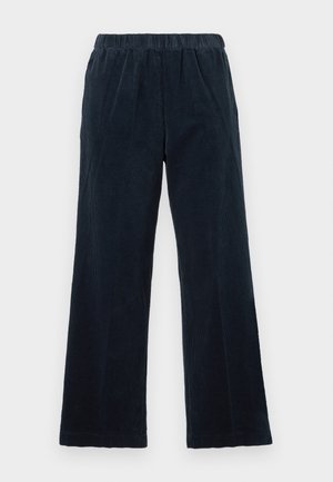 Nainen pukeutuneena mustaan pitkähihainen poolopaitaan, harmaaseen poolokaulukseen alla, tummiin leveälahkeisiin housuihin, mustiin slingback-pienikorkoisiin avokkaisiin ja pitää pientä mustaa olkalaukkua.
