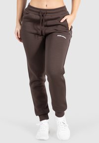 Frau trägt dunkelbraune Sweatpants mit Kordelzug und Reißverschlusstaschen sowie weiße Sneakers, steht vor einem hellgrauen Hintergrund.