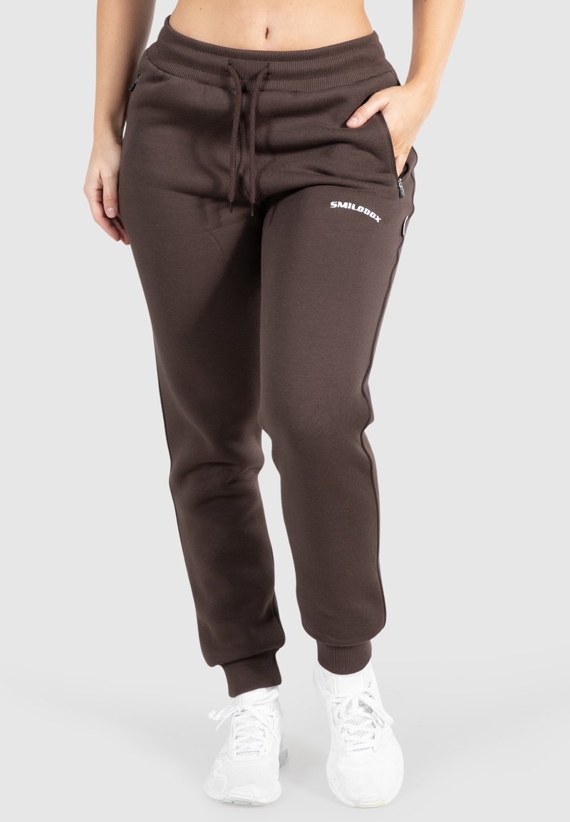 Frau trägt dunkelbraune Sweatpants mit Kordelzug und Reißverschlusstaschen sowie weiße Sneakers, steht vor einem hellgrauen Hintergrund.