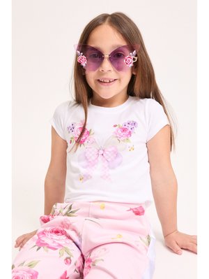 Fată tânără purtând ochelari de soare mov în formă de fluture, tricou alb cu imprimeu floral de fluturi și pantaloni roz cu flori, zâmbind așezată.