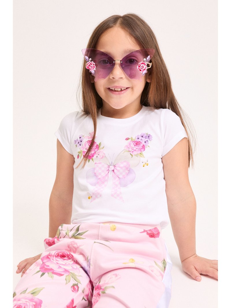 Νεαρή κοπέλα με μοβ γυαλιά ηλίου σε σχήμα πεταλούδας, λευκό πουκάμισο με παραστάσεις πεταλούδας και ροζ παντελόνι με λουλούδια, καθισμένη και χαμογελαστή.