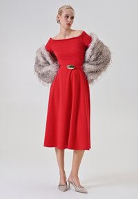 Rotes schulterfreies Kleid mit ausgestelltem Rock, tailliertem Bund und goldenem Gürtel. Akzentuiert mit einem flauschigen hellgrauen Schal und hellgrauen High Heels.