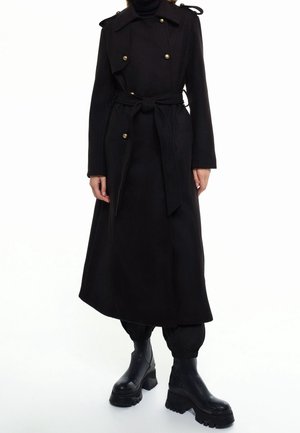 Mujer vestida con una gabardina larga negra de doble botonadura con botones dorados, atada a la cintura, pantalones negros y botas negras de plataforma.