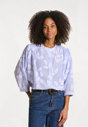 Blouse rayée bleu clair avec un motif de feuilles blanches, manches bouffantes et un devant boutonné. Associée à un jean en denim foncé et une ceinture noire.