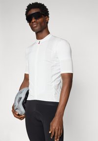 Maillot de cyclisme blanc à manches courtes, devant à demi-zip, avec un motif texturé subtil ; associé à un cuissard de cyclisme noir et un casque gris.