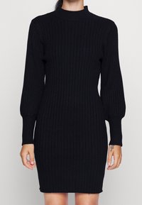 Abito nero a coste con colletto alto e maniche lunghe. L'abito presenta una silhouette aderente e un tessuto texturizzato in tutto.