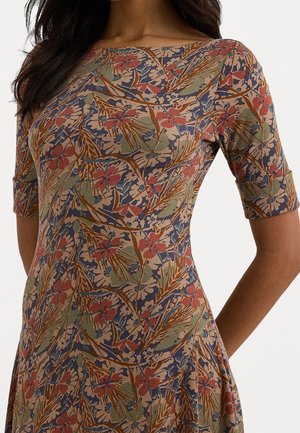 Frau trägt ein eng anliegendes Kleid mit kurzen Ärmeln und einem Blumenmuster in erdigen Tönen von Grün, Braun, Blau und Rot.