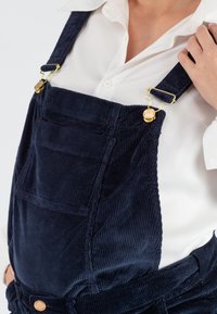 Marineblauwe corduroy salopettes met verstelbare bandjes hebben goudkleurige hardware en voorzakken. Draagt over een wit shirt, waarmee textuur en ontwerp worden benadrukt.